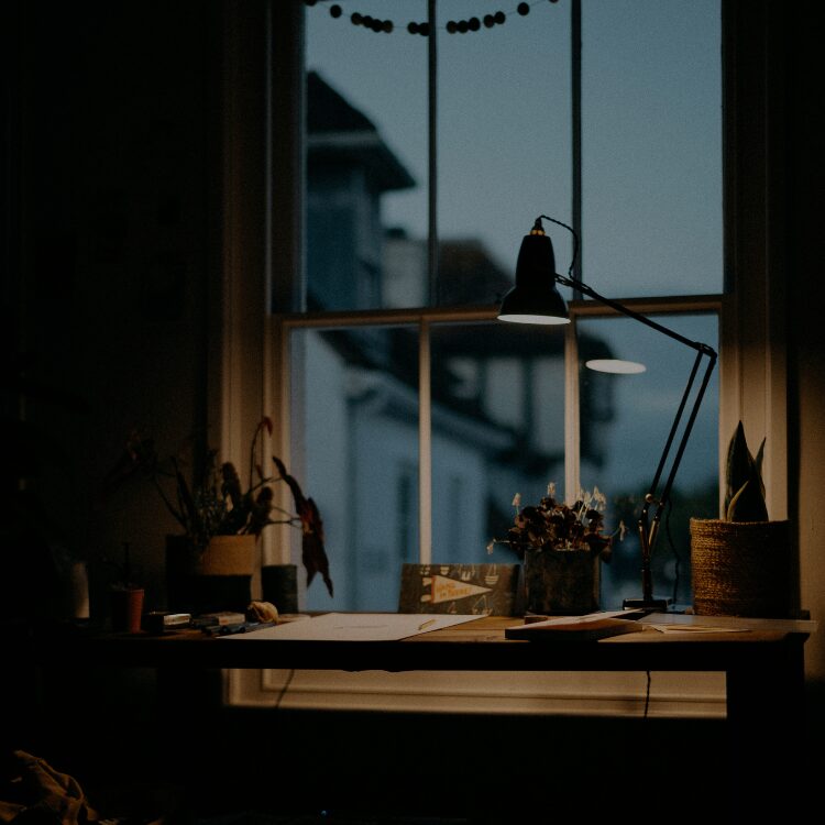 Desk by a large window at dusk with a black adjustable lamp, several potted plants, and scattered papers on the surface.