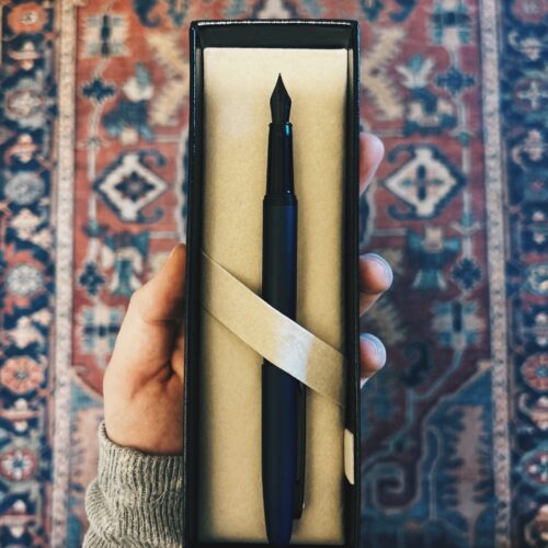 A black fountain pen sits in an open presentation box, held by a hand over a patterned rug background.