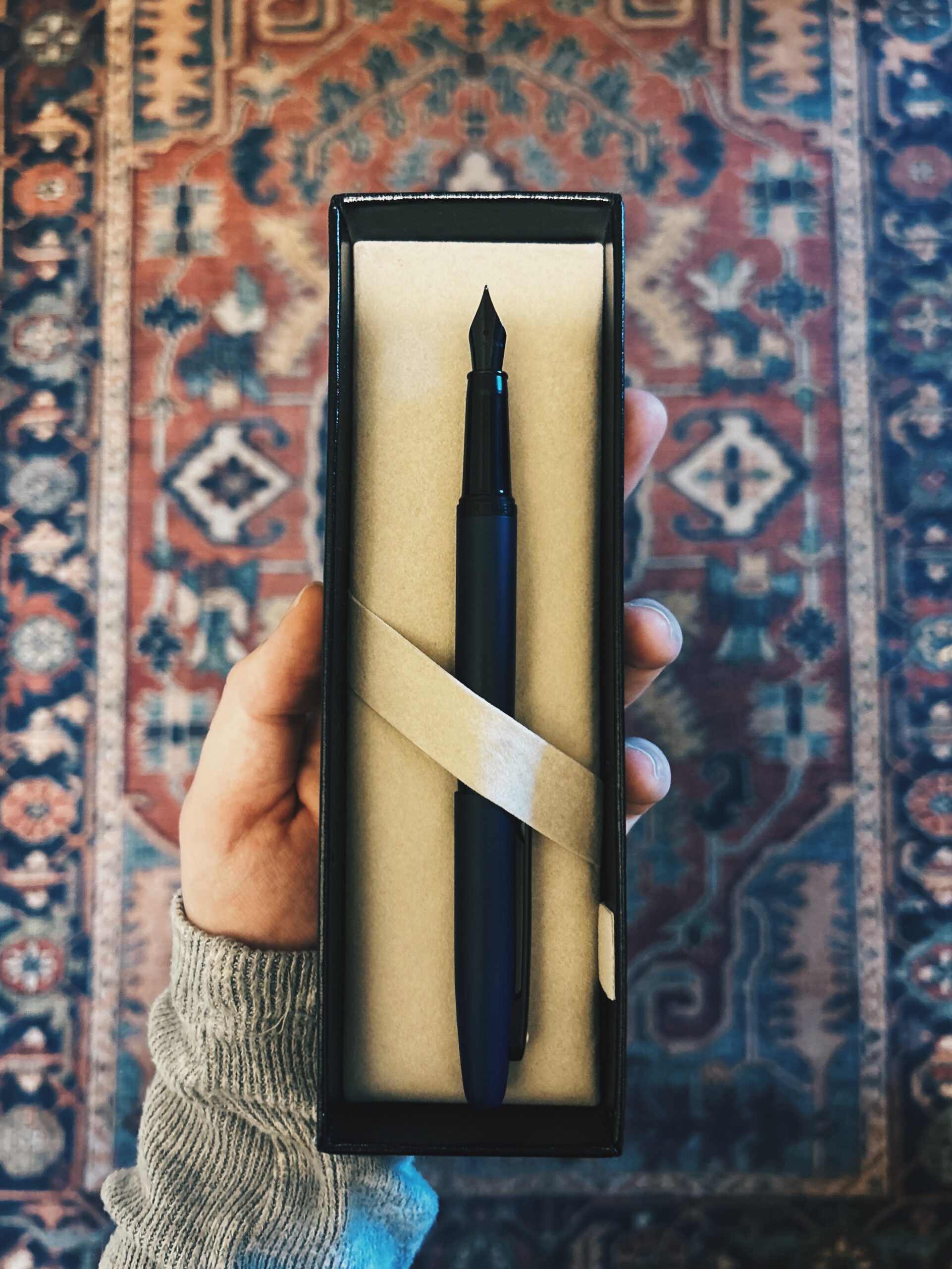 A black fountain pen sits in an open presentation box, held by a hand over a patterned rug background.
