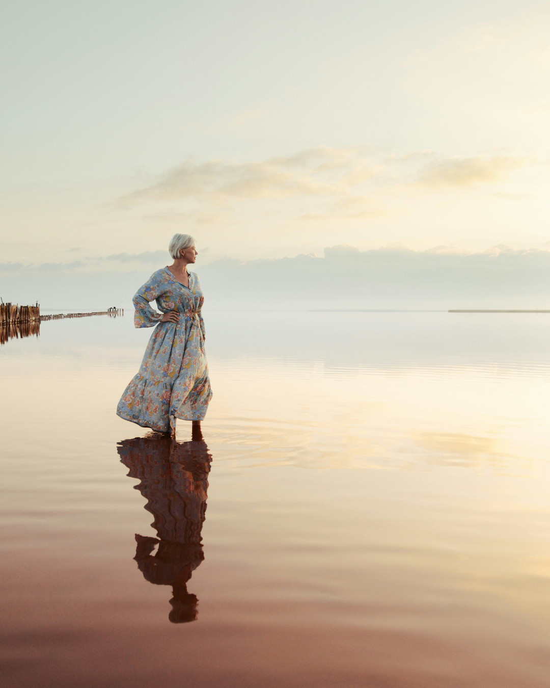 Older woman in a floral dress stands in shallow water at sunset, her reflection visible on the calm surface.