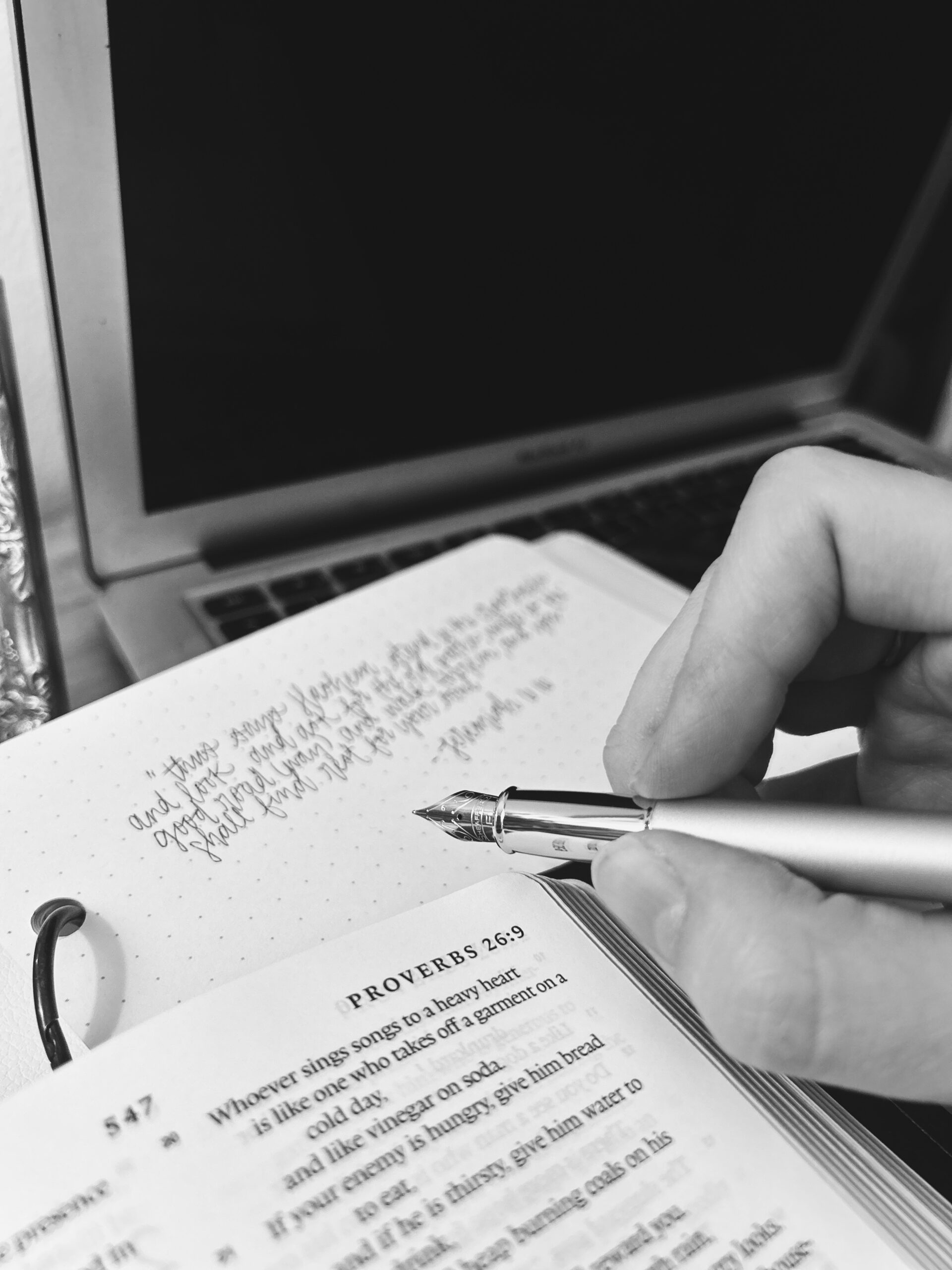 Close-up of a hand writing in a dotted notebook with a fountain pen; a laptop sits in the background and a Bible page shows Proverbs 26:9.