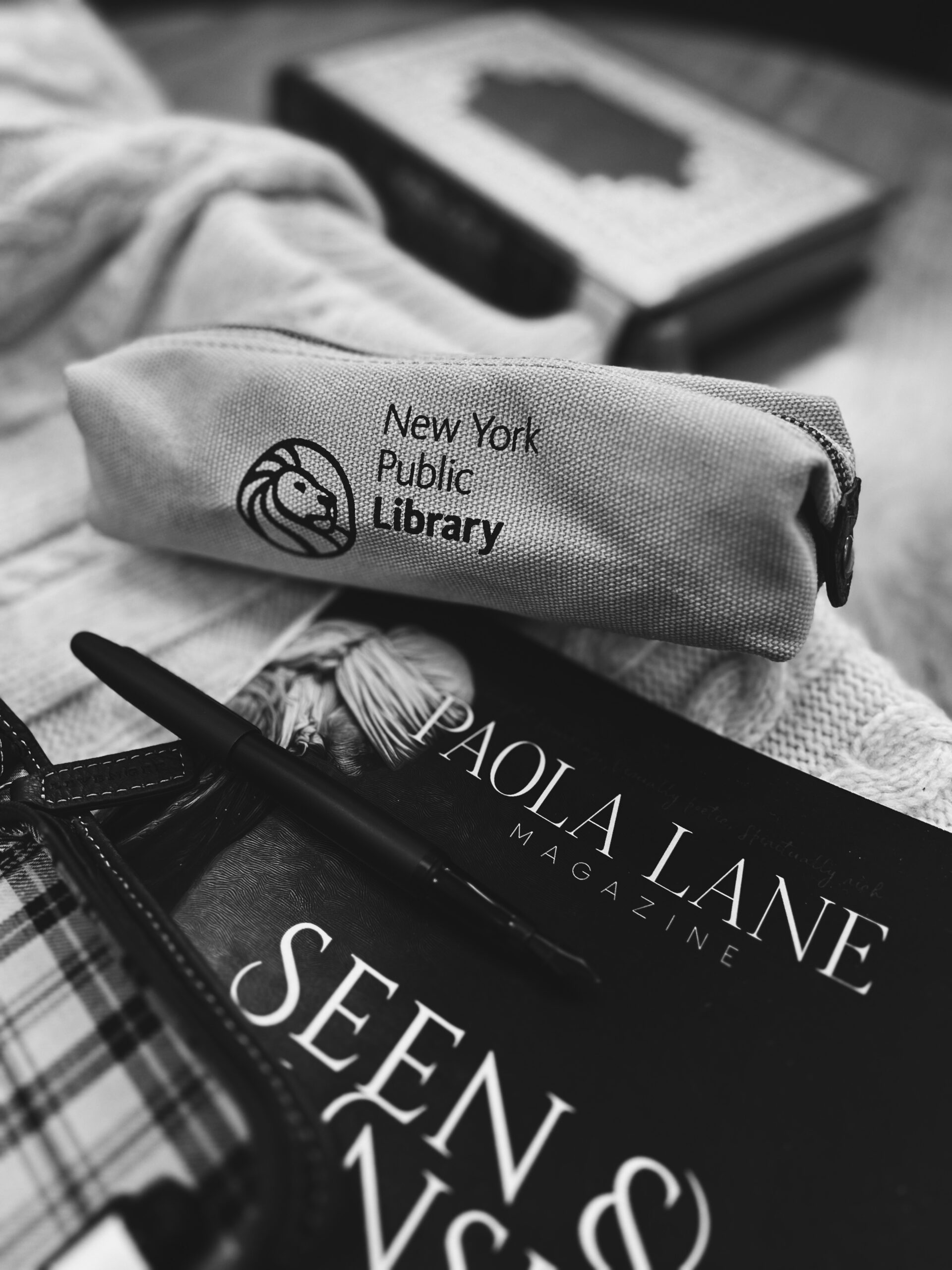 Close-up of a fabric pencil case displaying the New York Public Library logo on a desk behind stacked magazines.