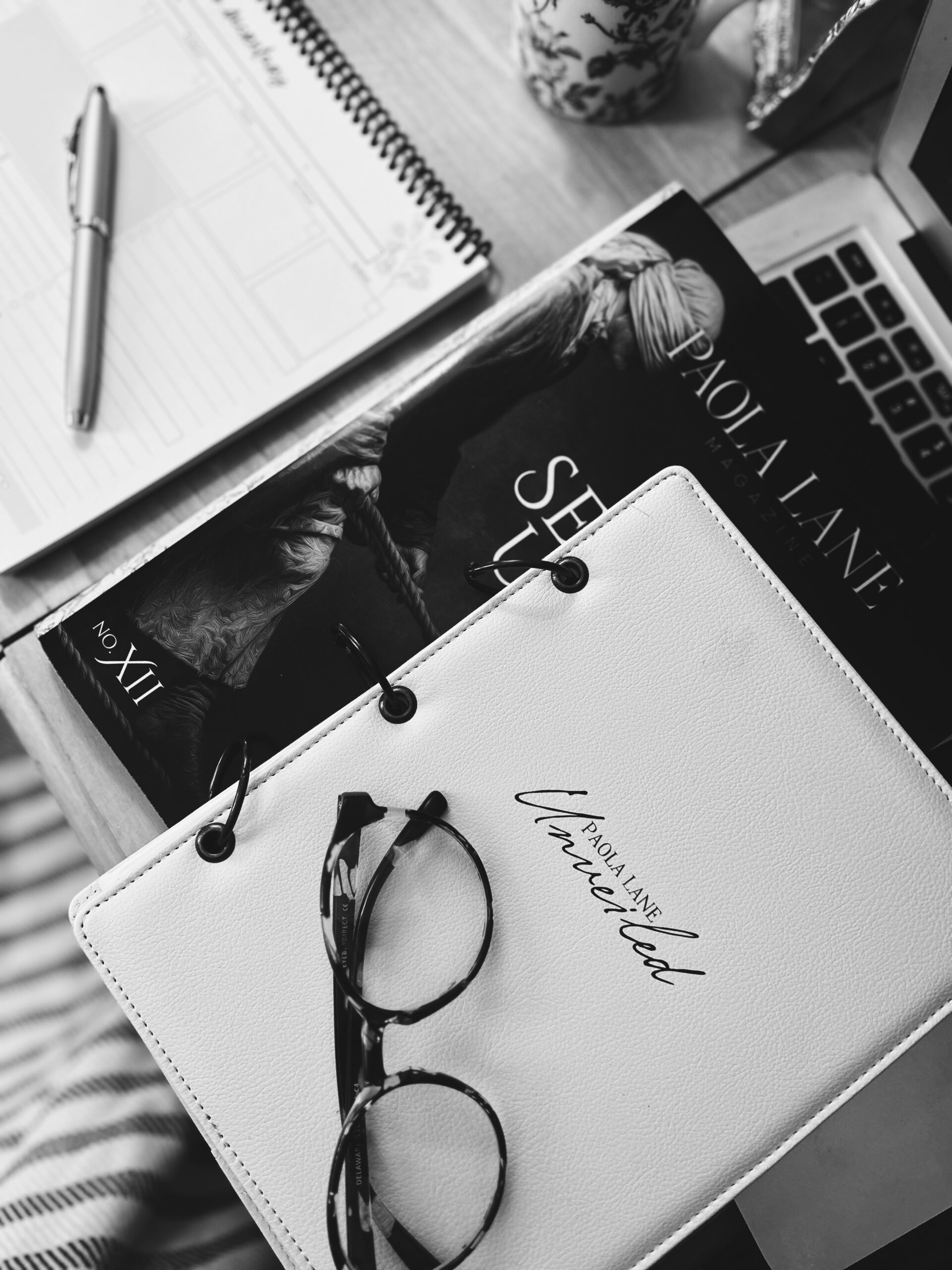 Black-and-white desk scene with a spiral notebook, pen, glossy magazine, and a pair of glasses resting on a white notebook near a laptop edge.