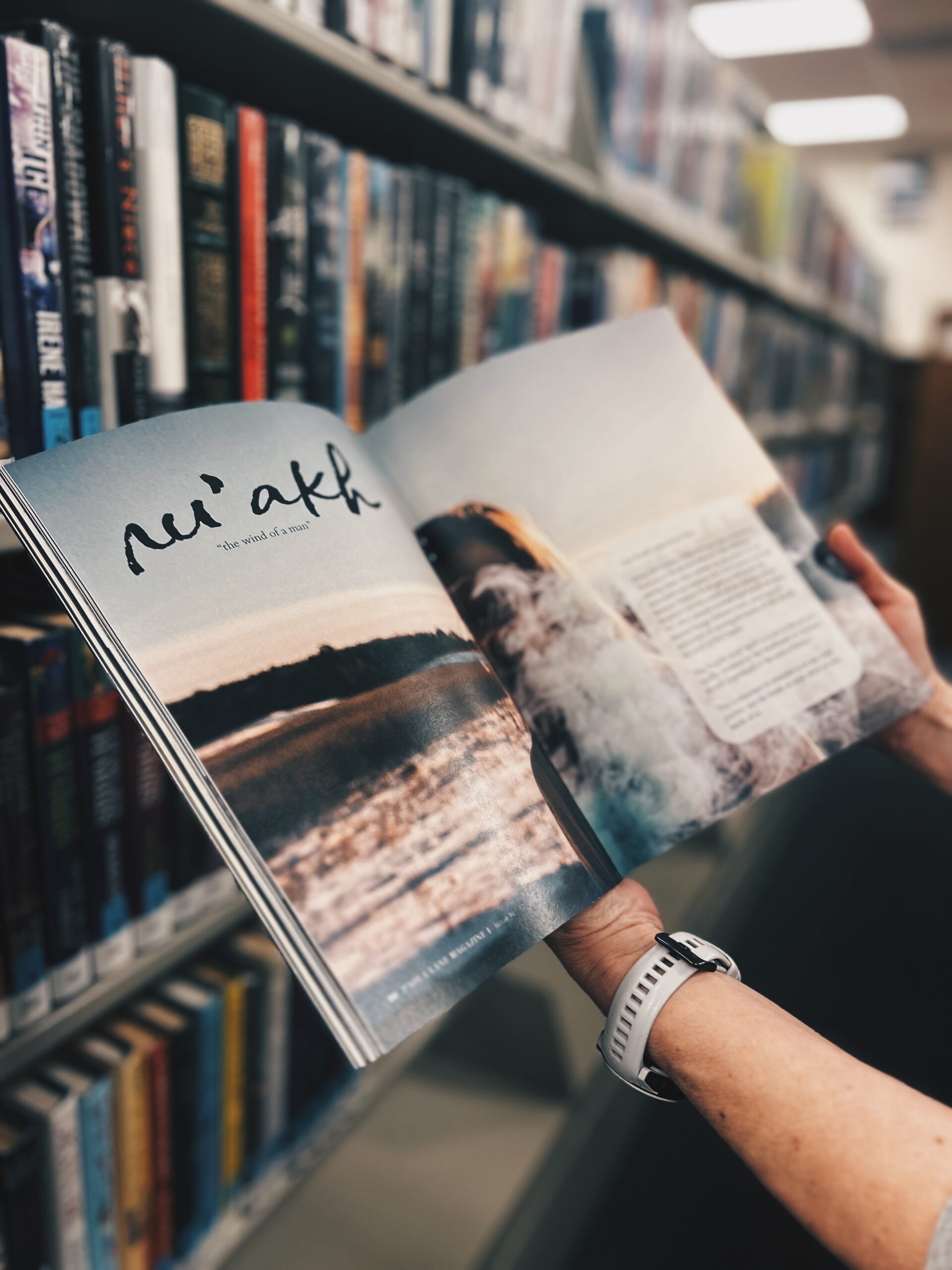 Person holding an open magazine with a landscape photo, in a bookstore or library aisle
