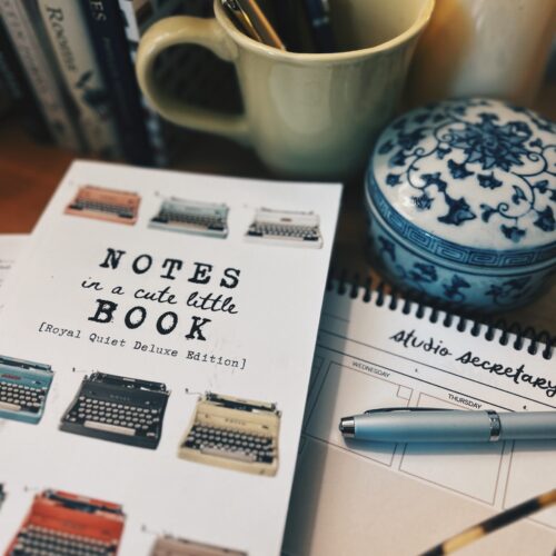 Beige mug with pens on a desk, next to a blue-and-white ceramic jar and a lit candle.