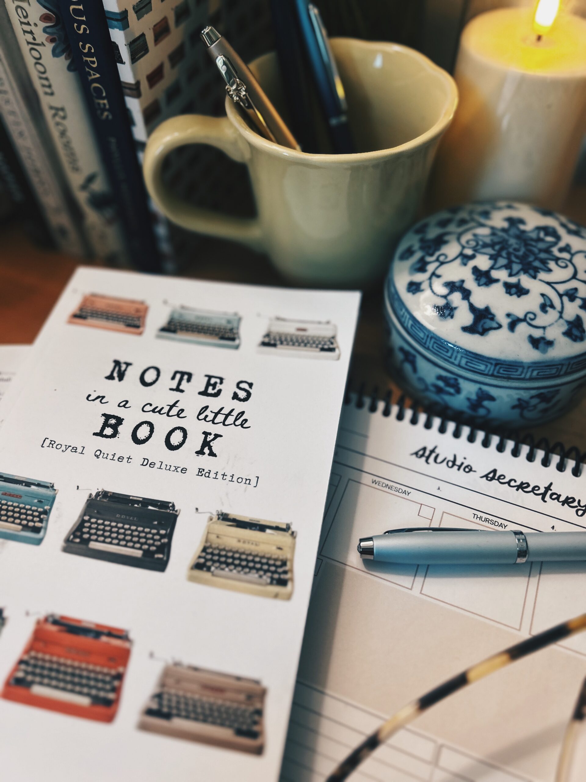Beige mug with pens on a desk, next to a blue-and-white ceramic jar and a lit candle.