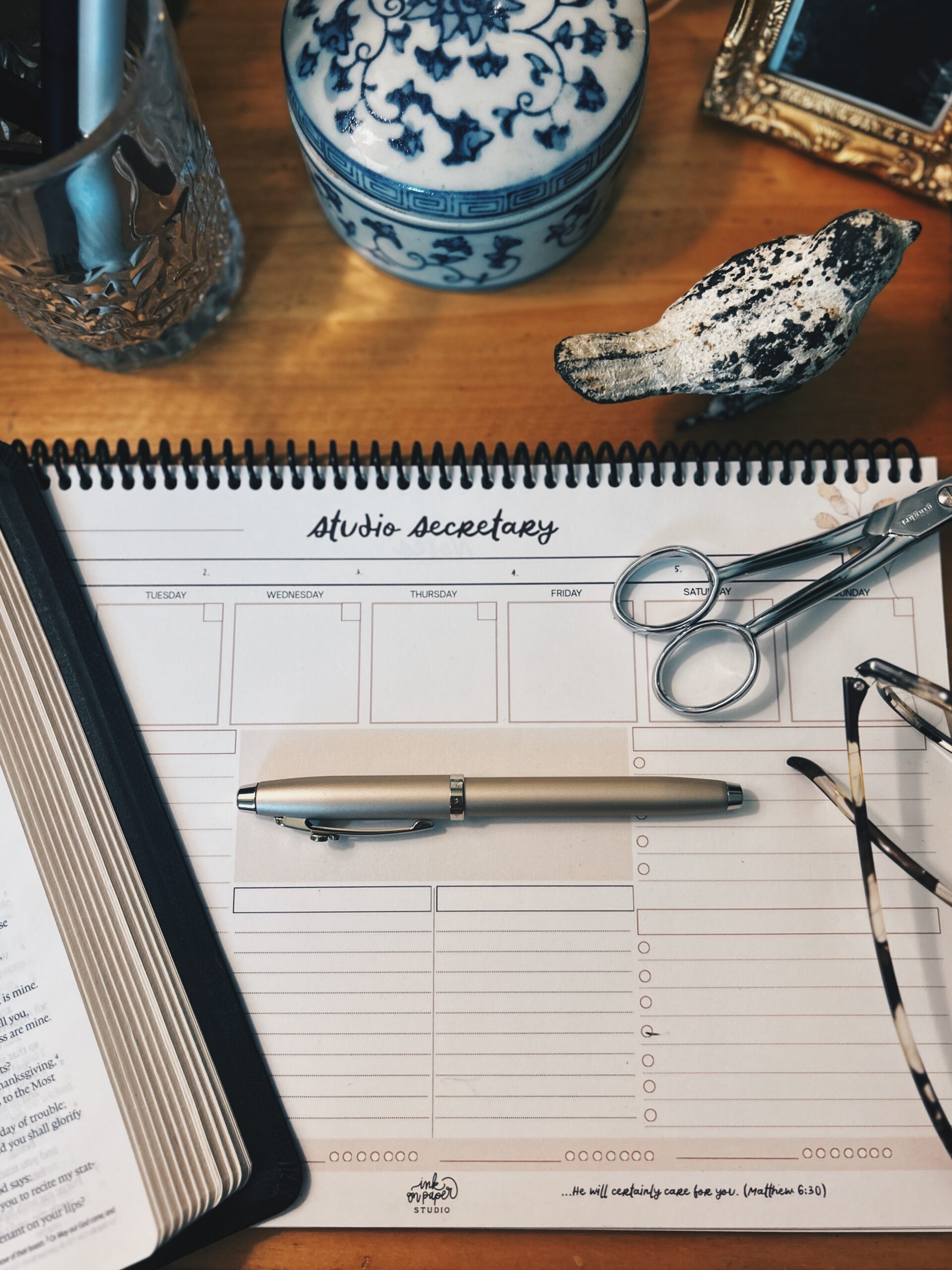 Top-down view of a wooden desk with a spiral-bound planner reading 'Studio Secretary', a silver pen, scissors, and glasses near a decorative blue-and-white ceramic container and a textured glass mug/place holder.