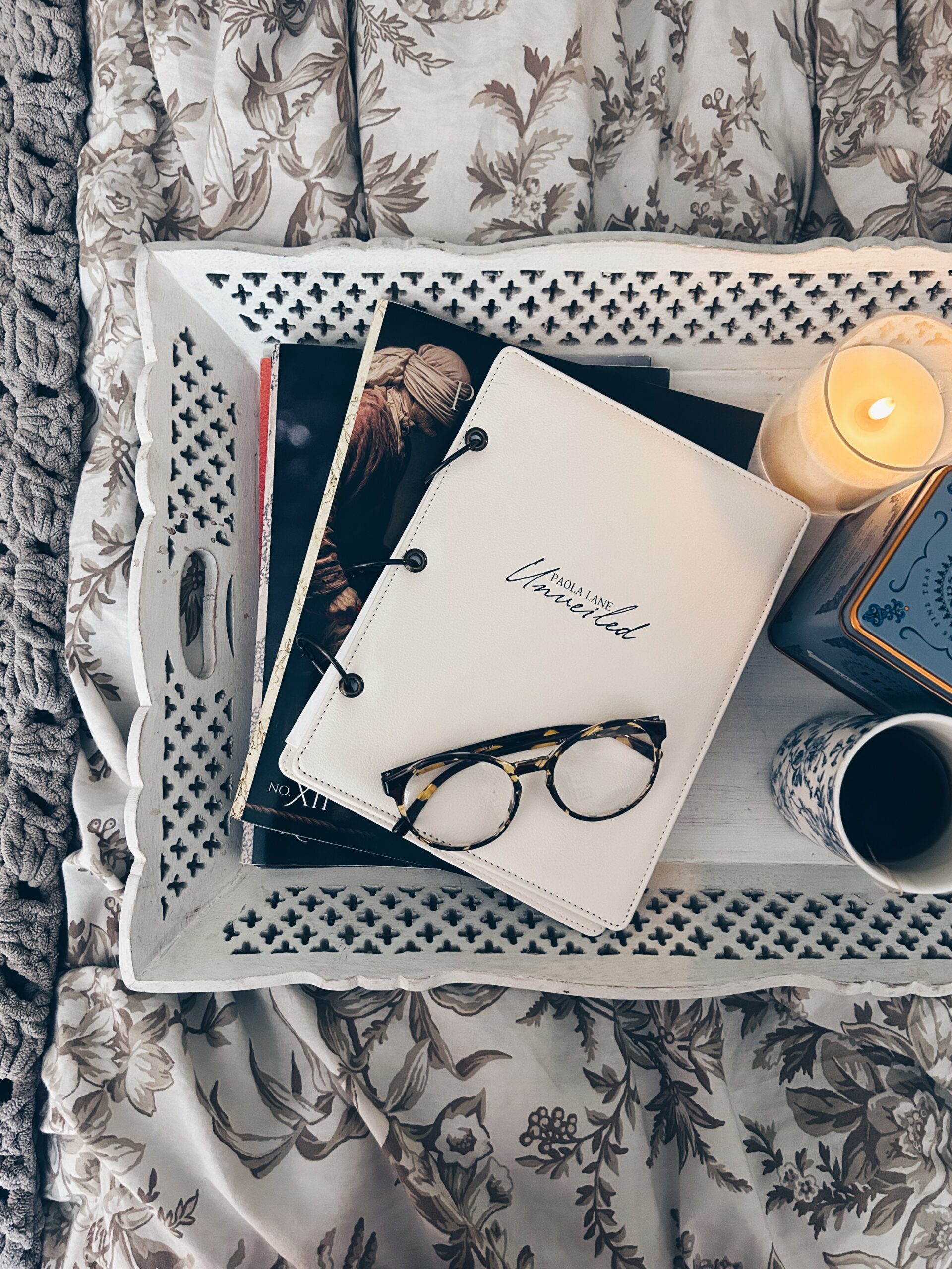 White decorative tray with cutout pattern holds a cream ring-bound notebook, some magazines, tortoiseshell glasses, and a lit candle on a floral bedspread.