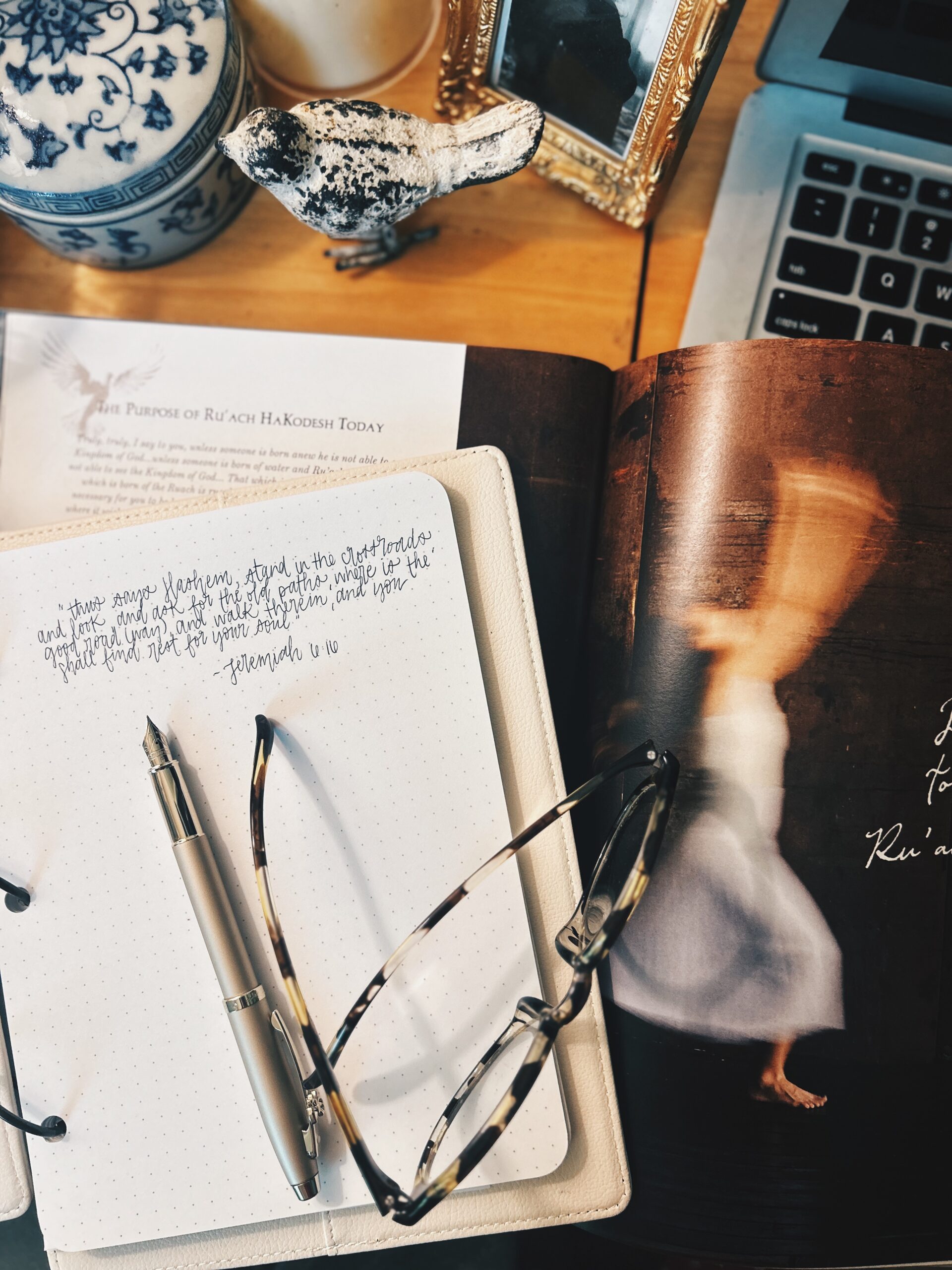 Desk study scene with an open dotted notebook, a beige pen and tortoiseshell glasses atop the pages, alongside a vinyl record and a laptop nearby.